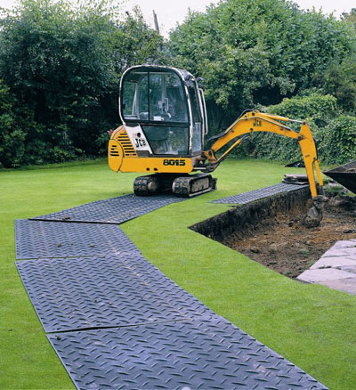 Road mats with a diamond tread pattern laid out on grass along the edge of a hole where a small digger is removing dirt from it